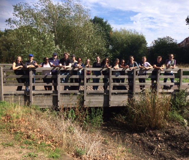 A large group of people standing on a wooden bridge facing the camera, they are smiling. In the background there are trees and there are some clouds in the sky.