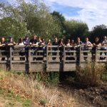 A large group of people standing on a wooden bridge facing the camera, they are smiling. In the background there are trees and there are some clouds in the sky.