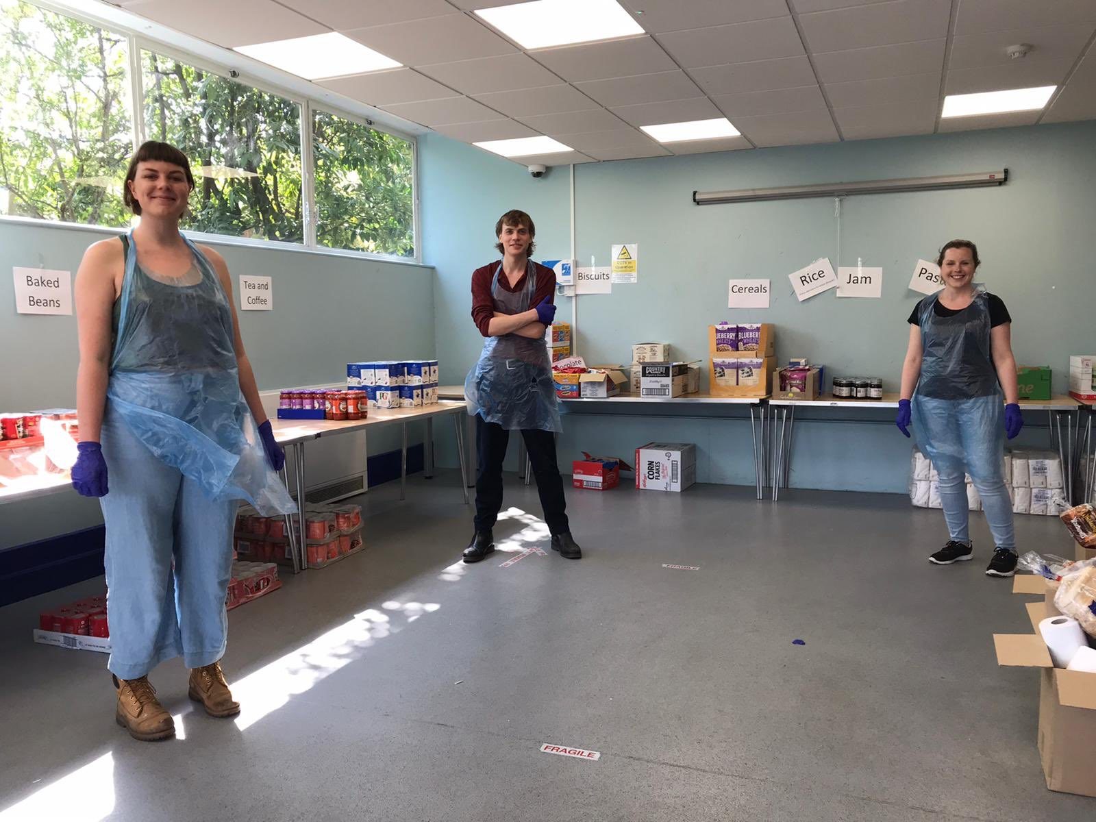 Three people are spaced out in a blue room, each wearing blue plastic aprons and blue gloves. There are tables along the walls of the room, covered with various food packaging.