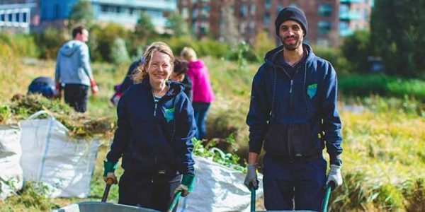 Two volunteers, a man and a woman, smiling while working outdoors in a green space. Both are holding wheelbarrows filled with vegetation. Other people in the background are seen gardening or clearing plants, with large bags of collected greenery nearby. The scene is set against an urban backdrop with buildings and greenery.